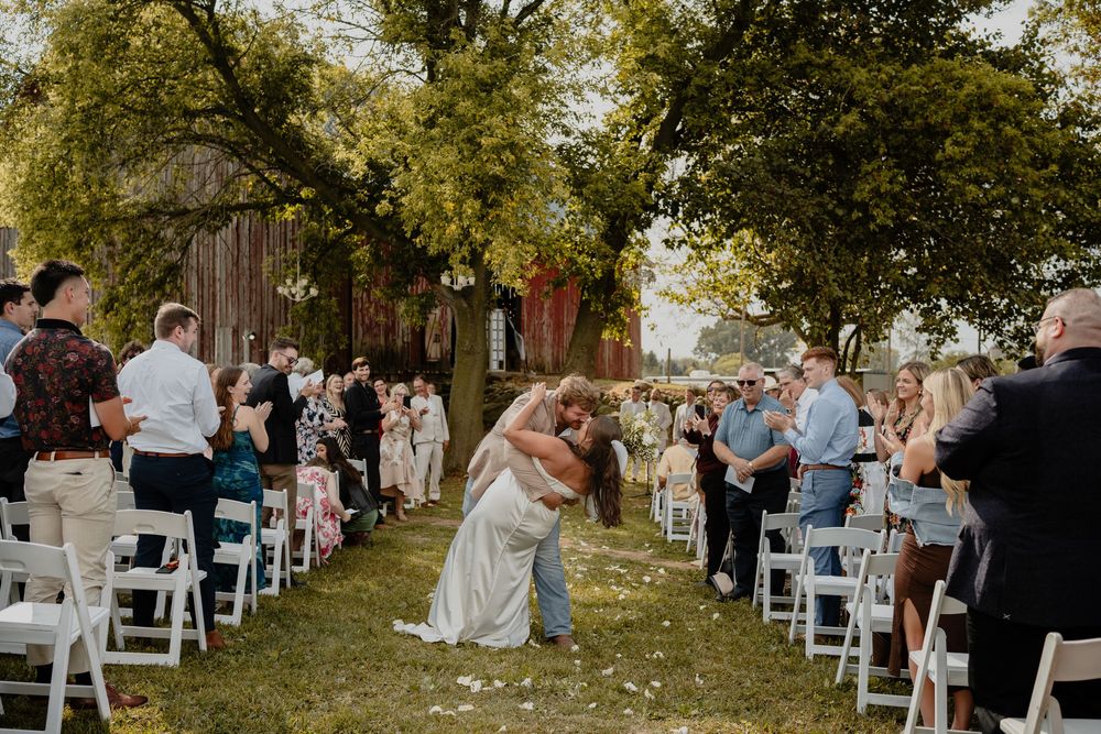 Wedding guests gather outdoors under a canopy of trees during an autumn ceremony with white chairs arranged in rows.