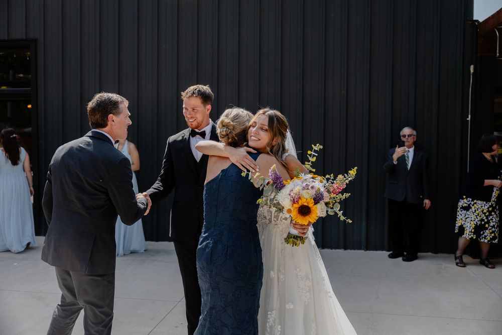 Bride in white gown holding sunflower bouquet embraces guest at outdoor wedding reception near dark barn.