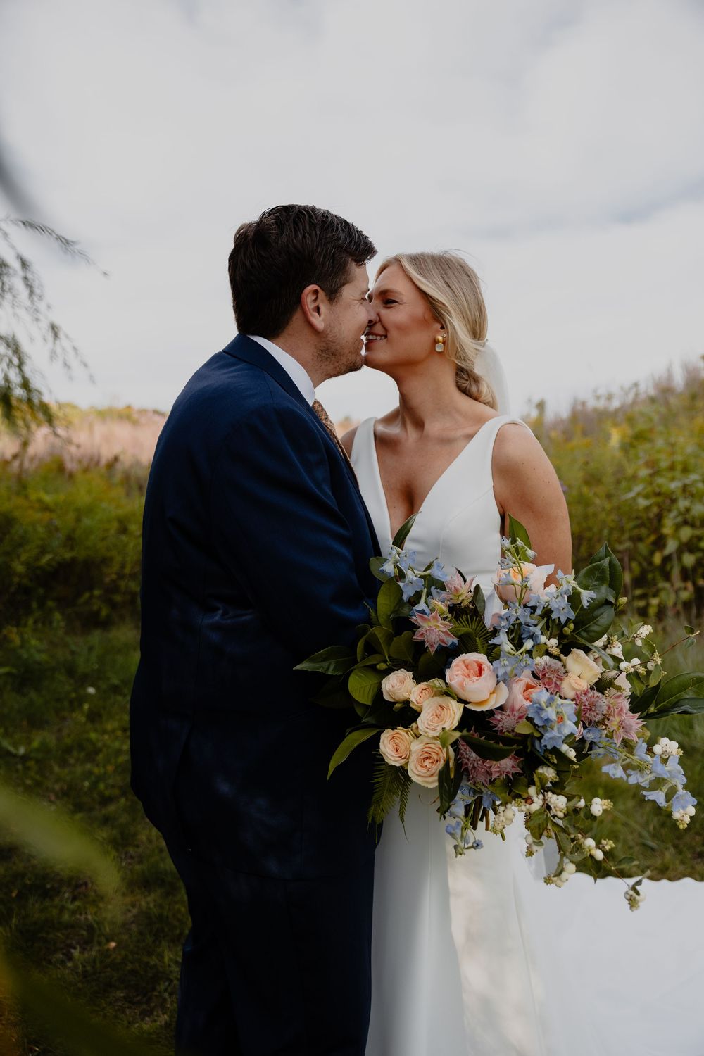 A romantic wedding couple shares a tender kiss while holding a lush bouquet of peach and blue flowers outdoors.