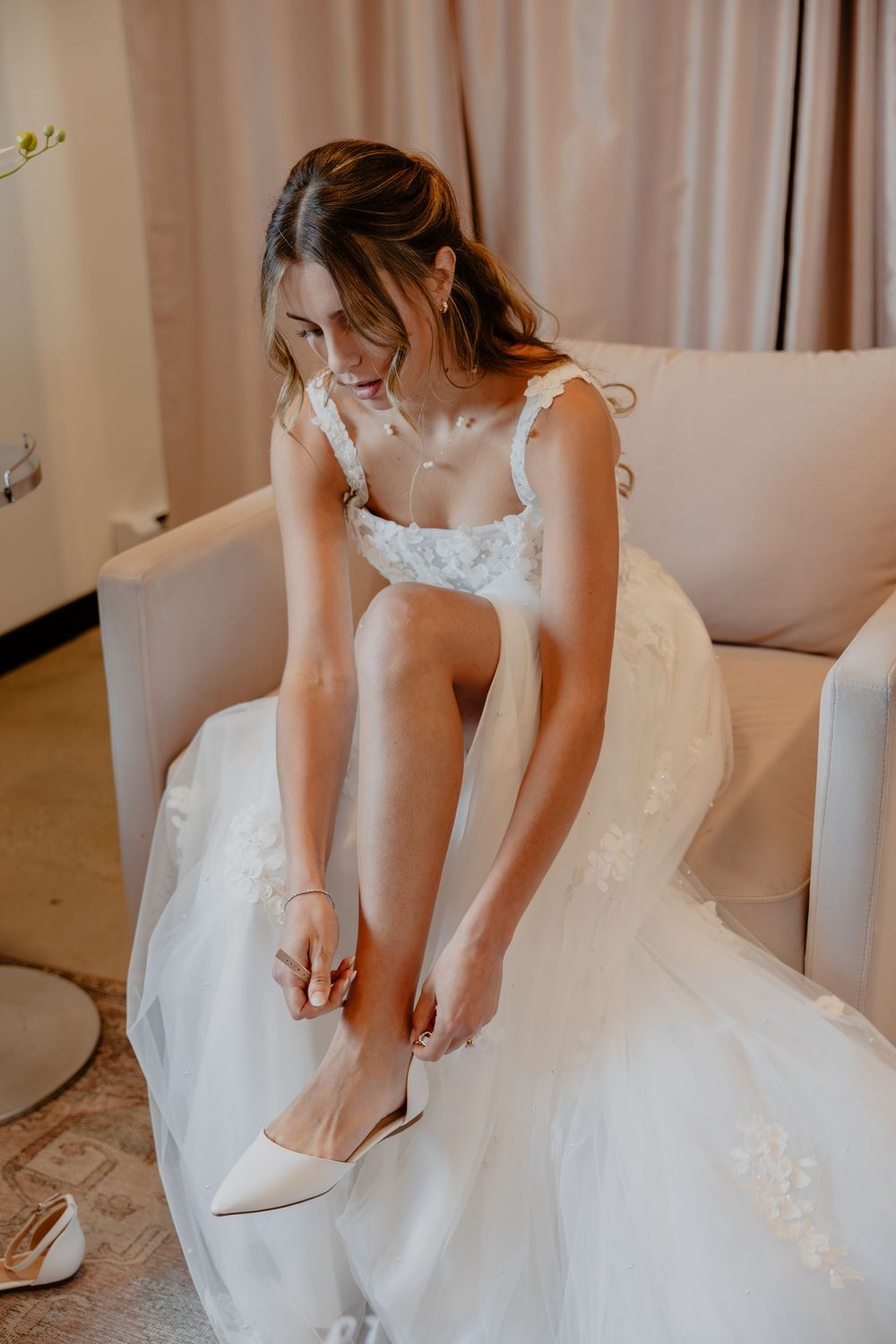 Bride in white lace wedding gown sitting on cream chair putting on ivory heels while getting ready.
