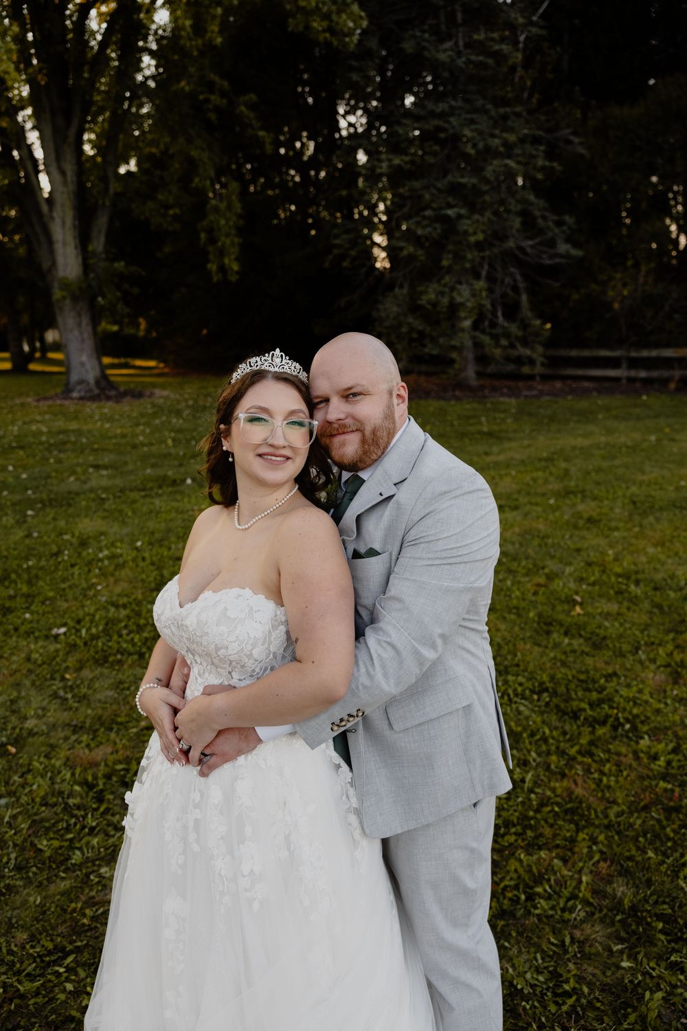 A couple in wedding attire embrace outdoors in a grassy park setting during a romantic photoshoot.