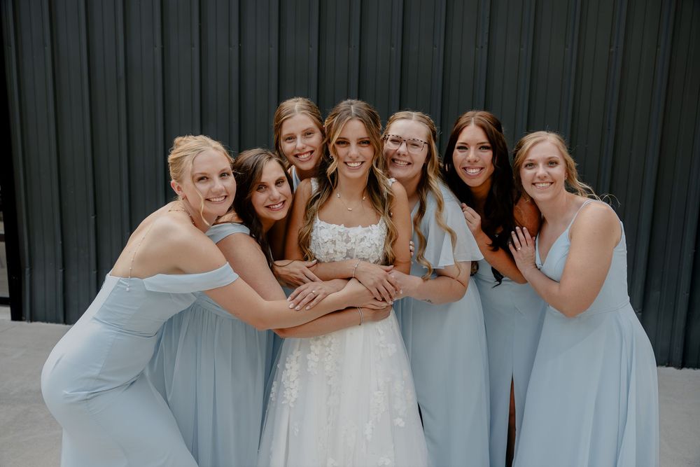 Bride in white lace gown surrounded by six bridesmaids in light blue dresses, smiling together indoors.