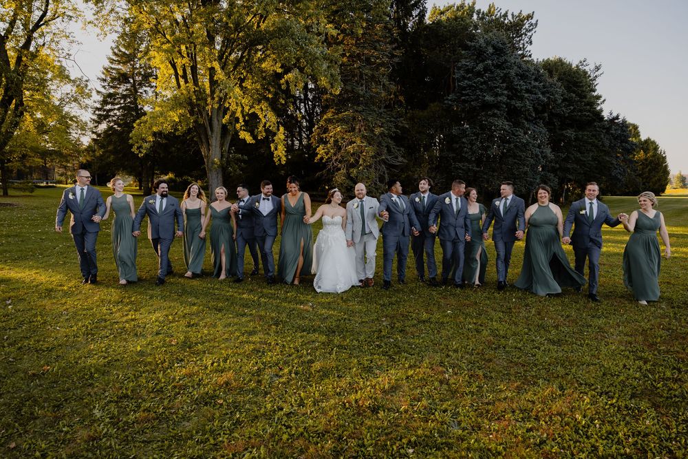Wedding party stands in a line on a grassy hillside during golden hour at an autumn outdoor celebration.