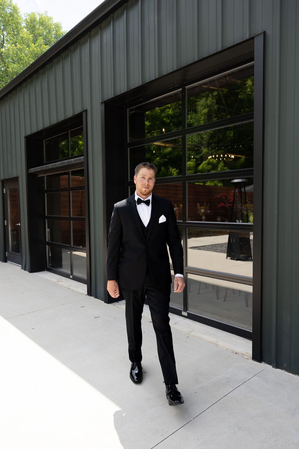 Groom in black tuxedo with bow tie and white pocket square standing outside modern green building.
