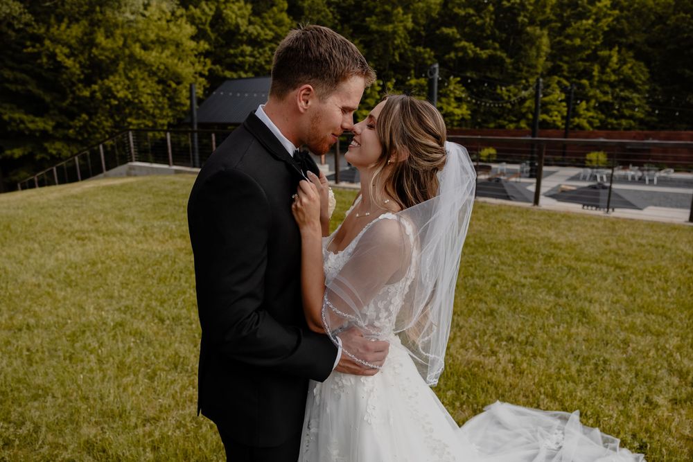 Bride and groom share a romantic almost-kiss outdoors, dressed in wedding attire with a veil and black tuxedo.