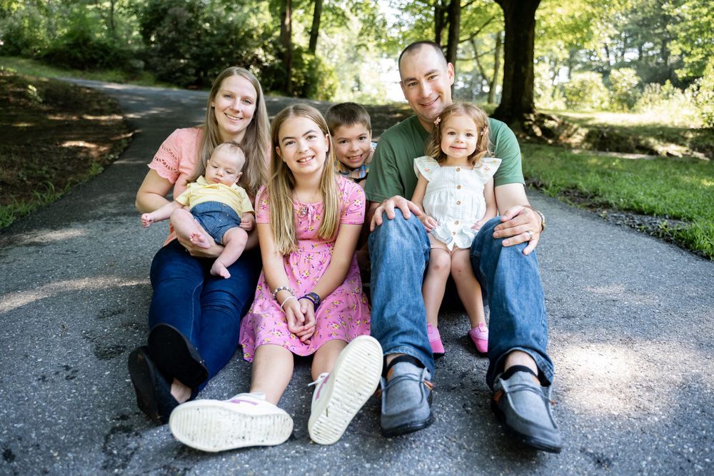 A family group sits together outdoors on a paved pathway surrounded by greenery.