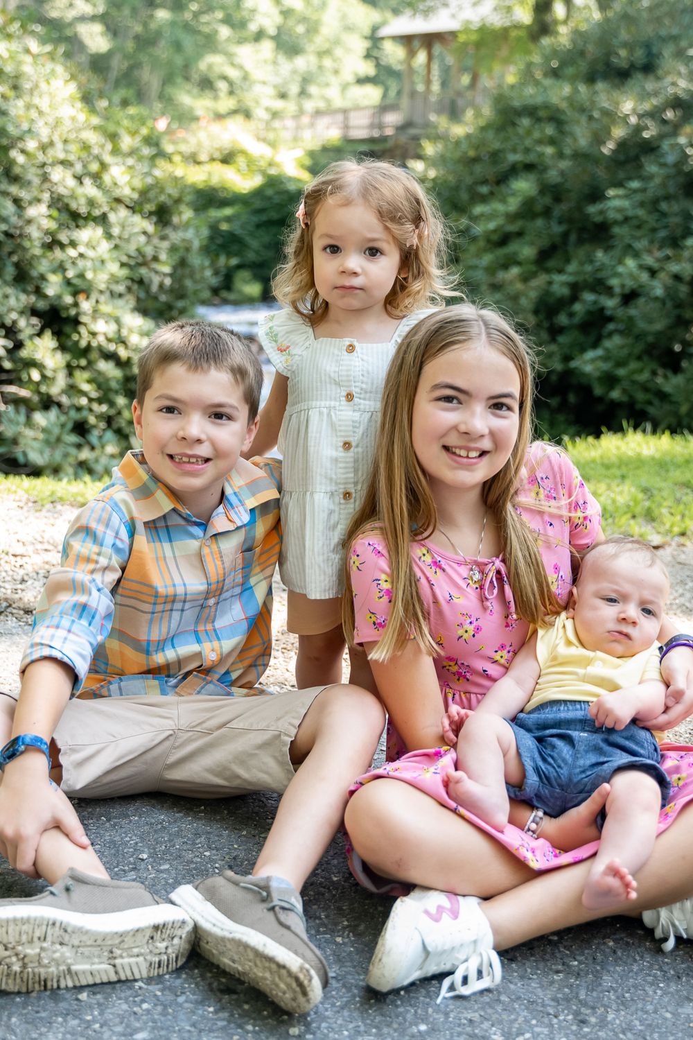 Four children sit casually together on a paved outdoor path with trees in the background.