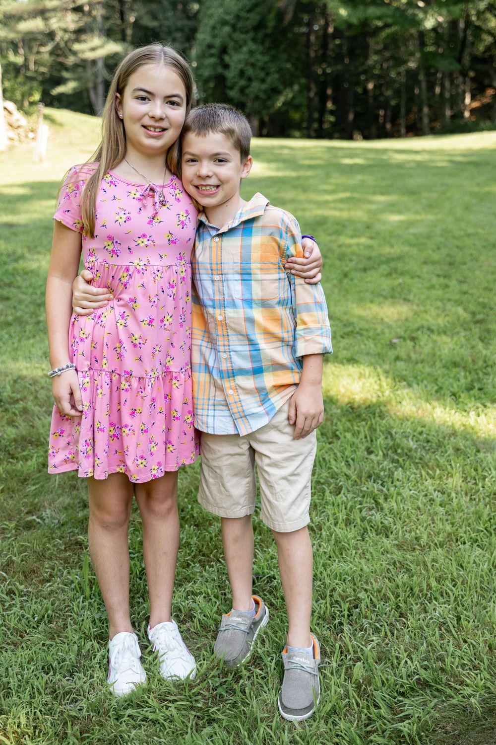 Portrait of two siblings in summer outfits standing together on sunny lawn.
