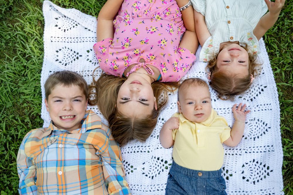 Four children lying on white lace blanket in grass forming circle pattern.