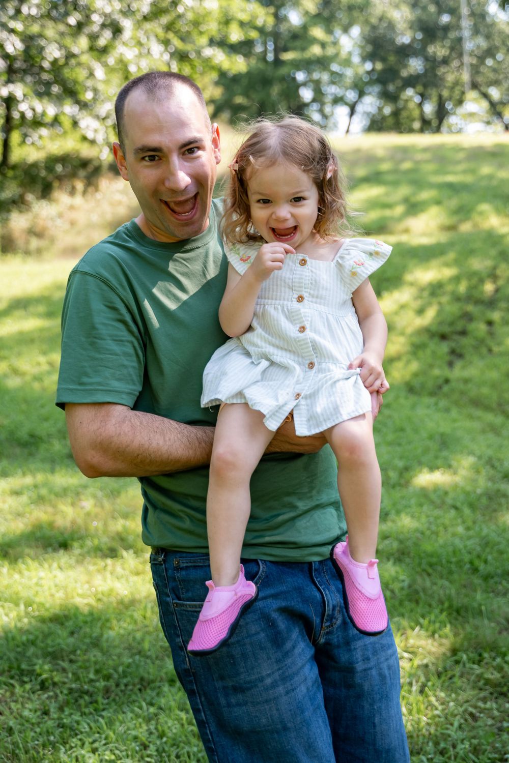 A joyful outdoor portrait shows a small child in a white dress and pink shoes being held on a sunny grassy hill.