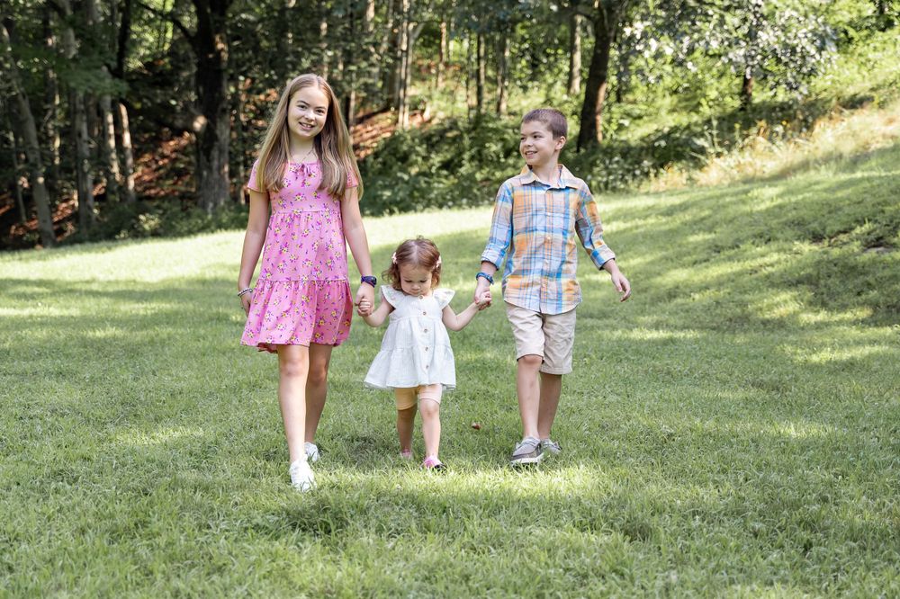 Three children walking together holding hands on sunny grassy hillside.