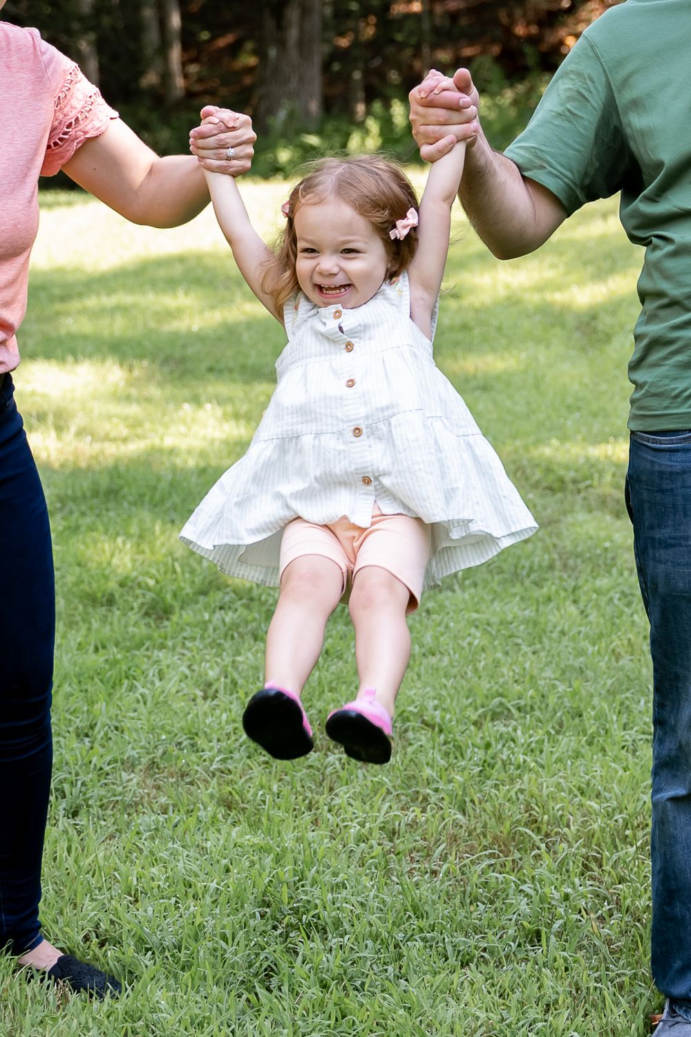 A child in a white dress swings playfully between two adults on a sunny lawn.