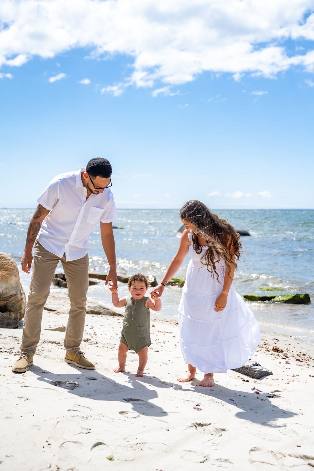 A family holds hands while walking together on a sunny beach with ocean waves in the background.