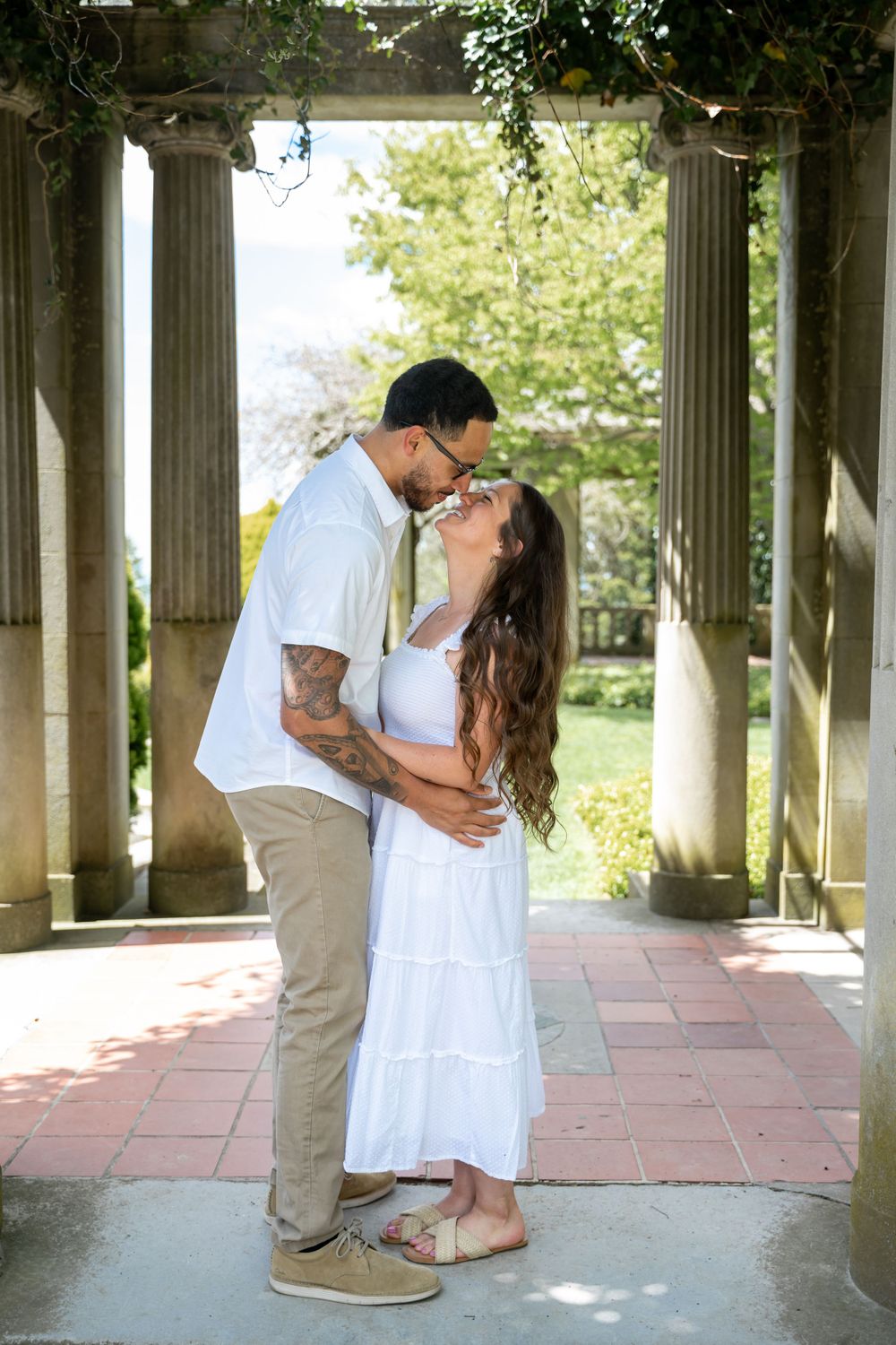 Romantic couple embracing under a columned pergola surrounded by greenery.