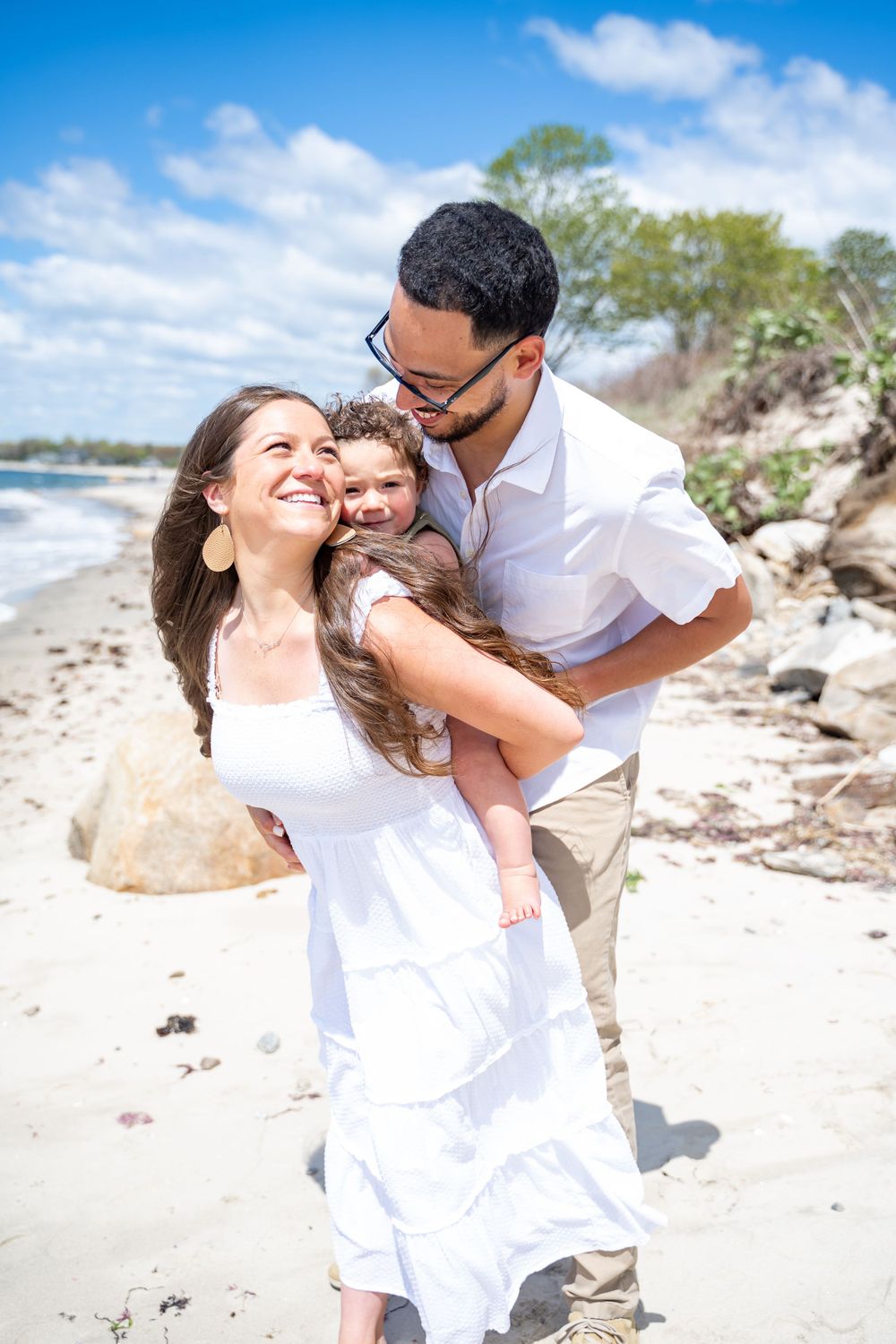 A family embraces while enjoying a sunny day at the beach with rocky coastline in the background.