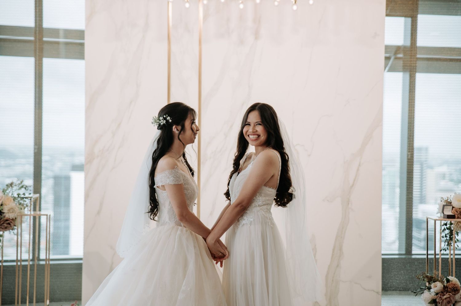 Two people in white wedding dresses holding hands against a pink backdrop.