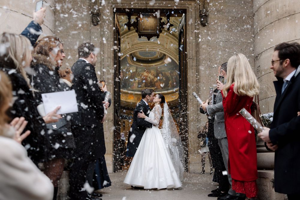 Wedding couple exits historic doorway as guests shower them with confetti in celebration.