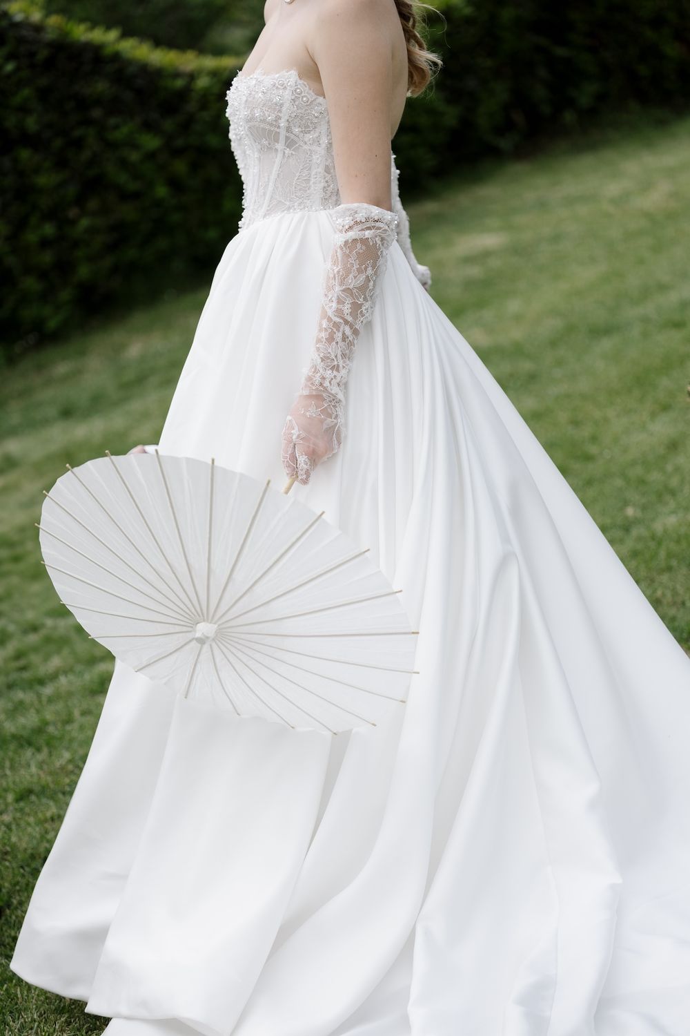 Elegant bride in flowing white gown holds decorative paper parasol in garden setting.
