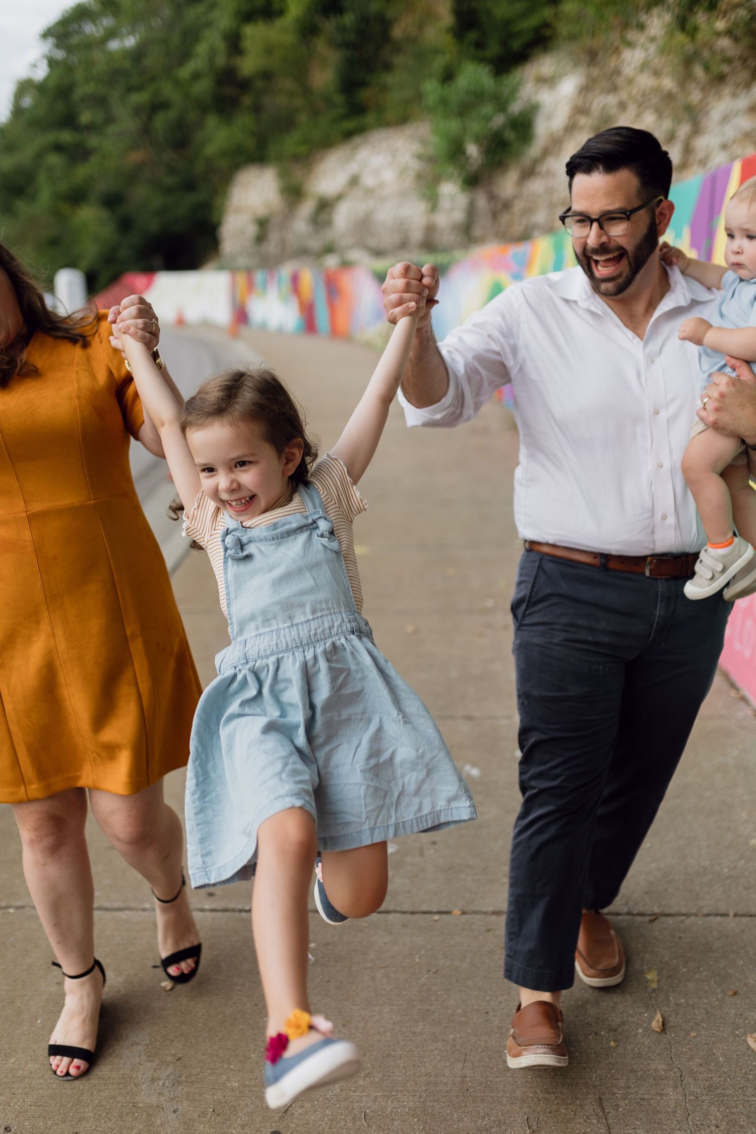 A family swings their young daughter in the air candidly as she laughs while walking in front of the Roeland Park mural.