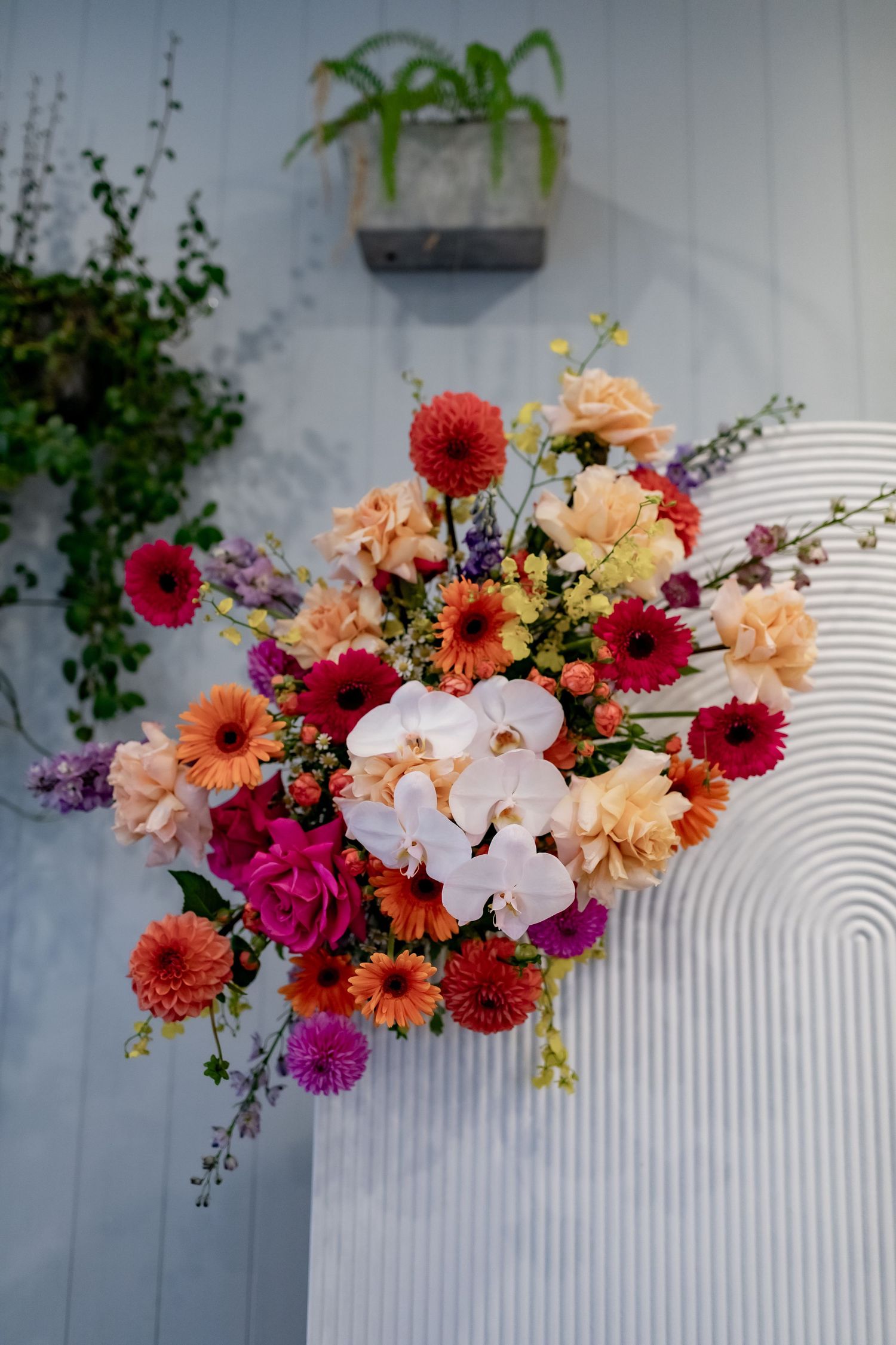 Vibrant floral arrangement with pink, orange, and white flowers against a textured white wall.