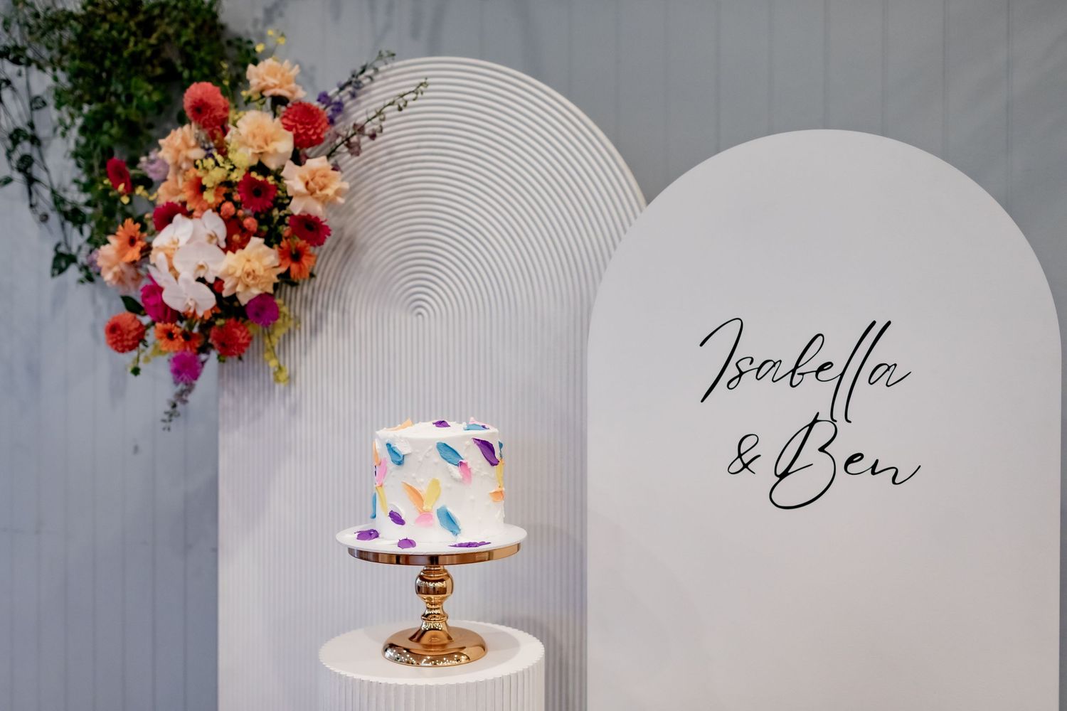 Modern wedding cake display with colorful floral arrangement against white curved backdrop.