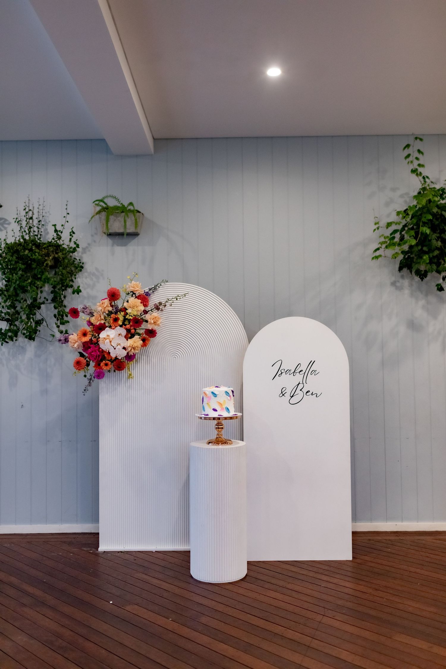 Modern event backdrop with white arches, floral arrangement and decorative cake display.