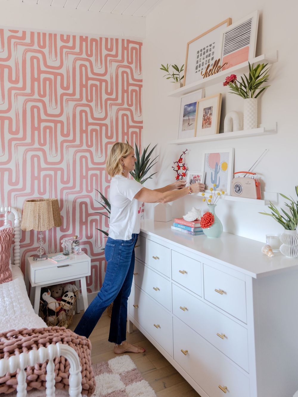 Interior designer arranging decor on white dresser against pink geometric wallpaper in modern nursery.