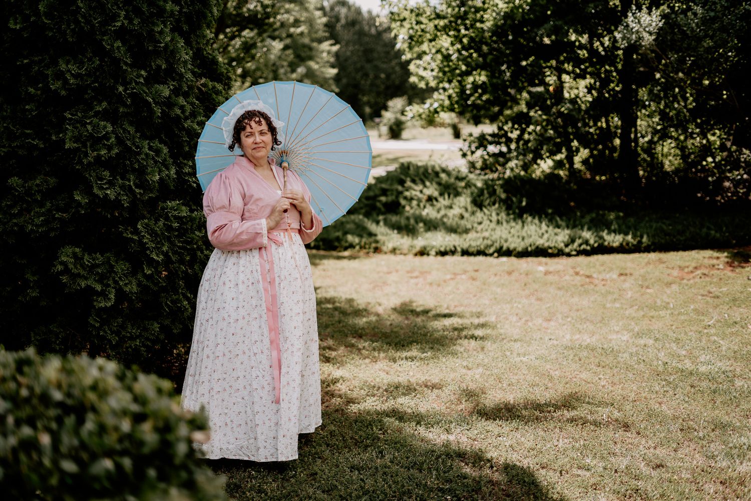 A person in a pink vintage dress holds a light blue parasol in a sunny garden setting.