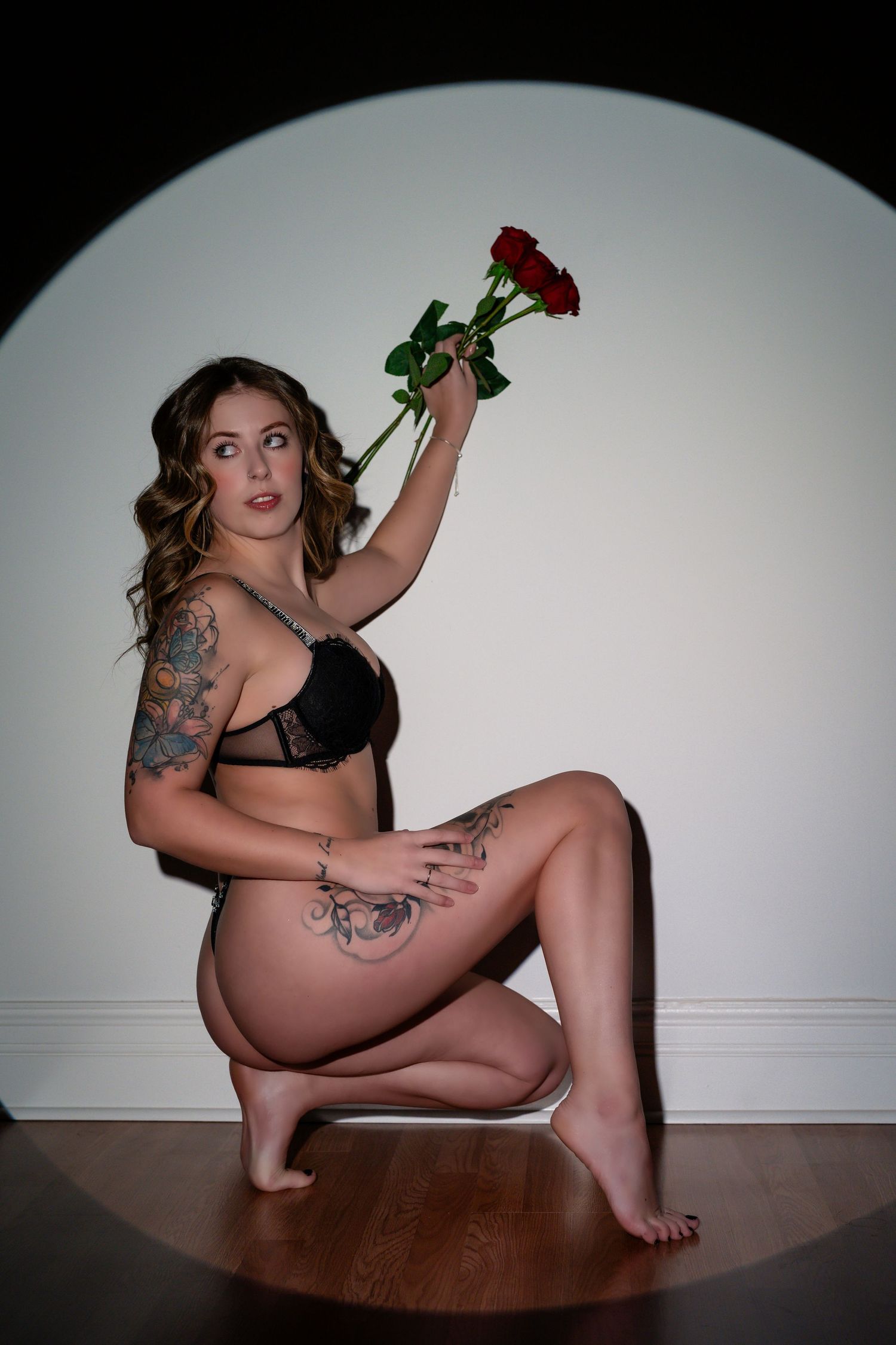 Playful boudoir pose holding red rose against white circular backdrop.