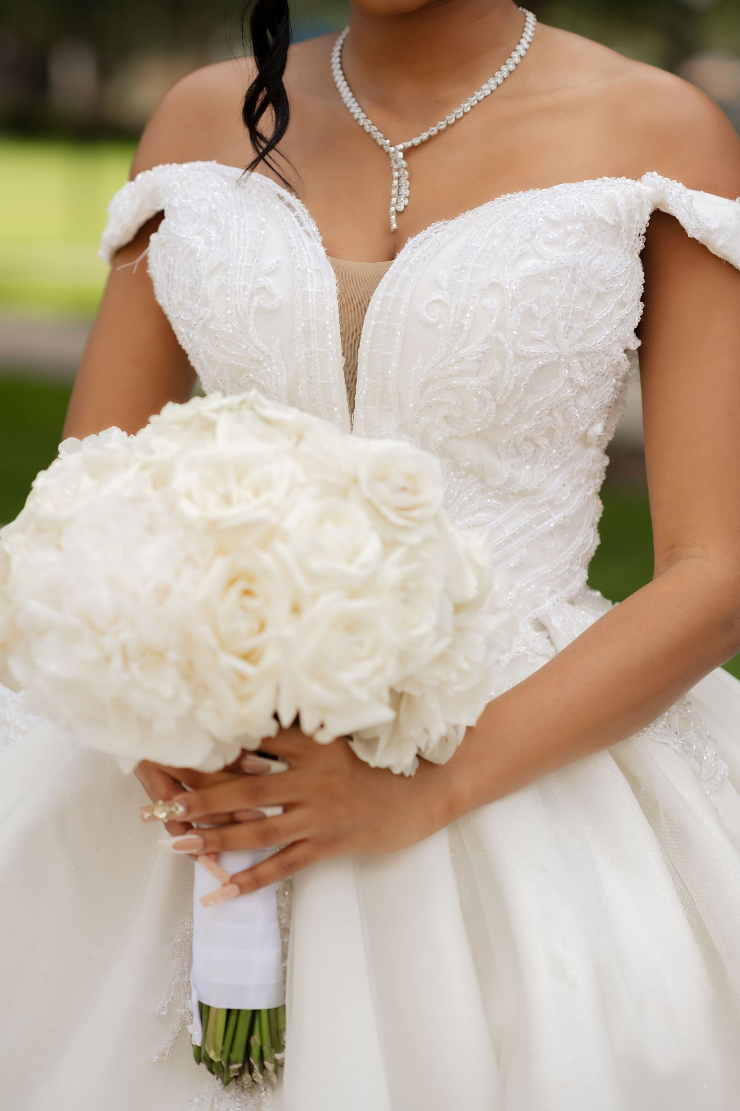 Elegant bridal detail showing white off-shoulder dress, diamond necklace and white rose bouquet.