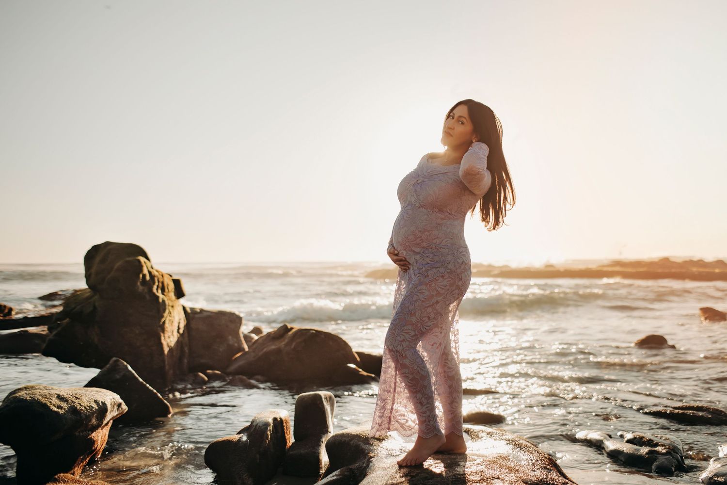 Maternity session with a woman in a purple dress, standing on a rock at the beach, holding her hair, capturing a serene and dramatic moment.