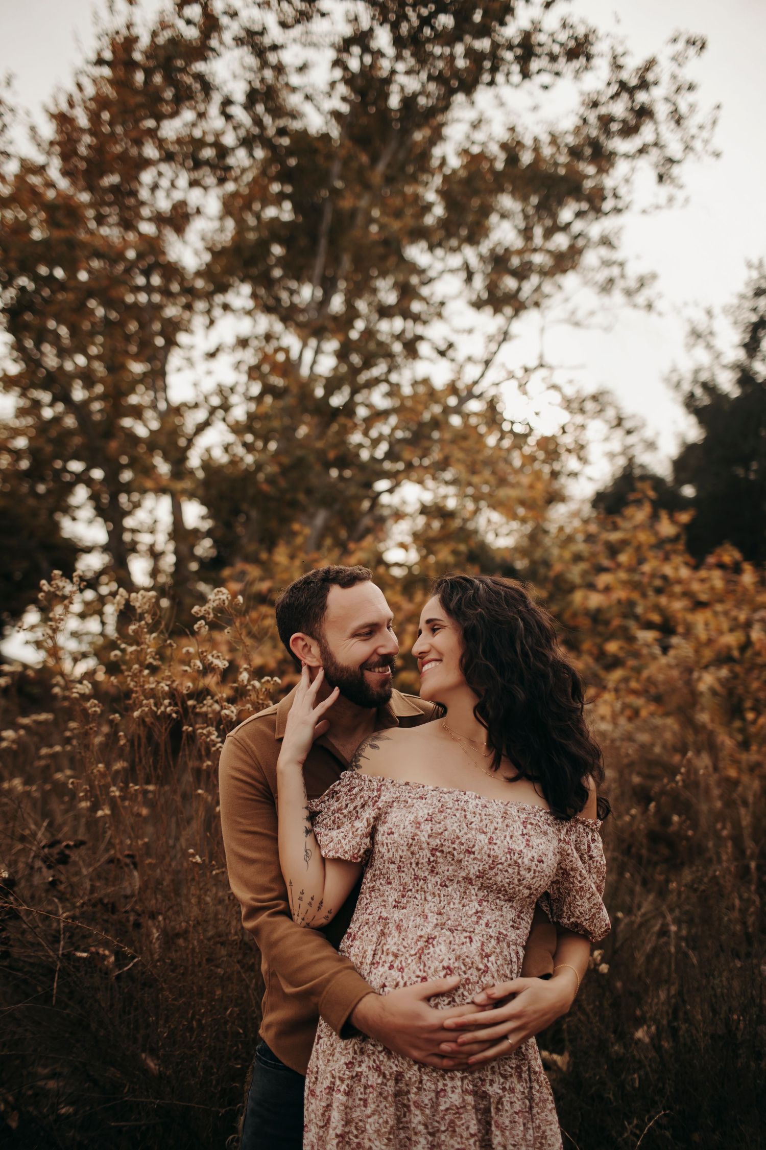 Maternity session in a setting with fall leaves, the husband holding his wife's belly as she looks at him lovingly, sharing a tender moment.