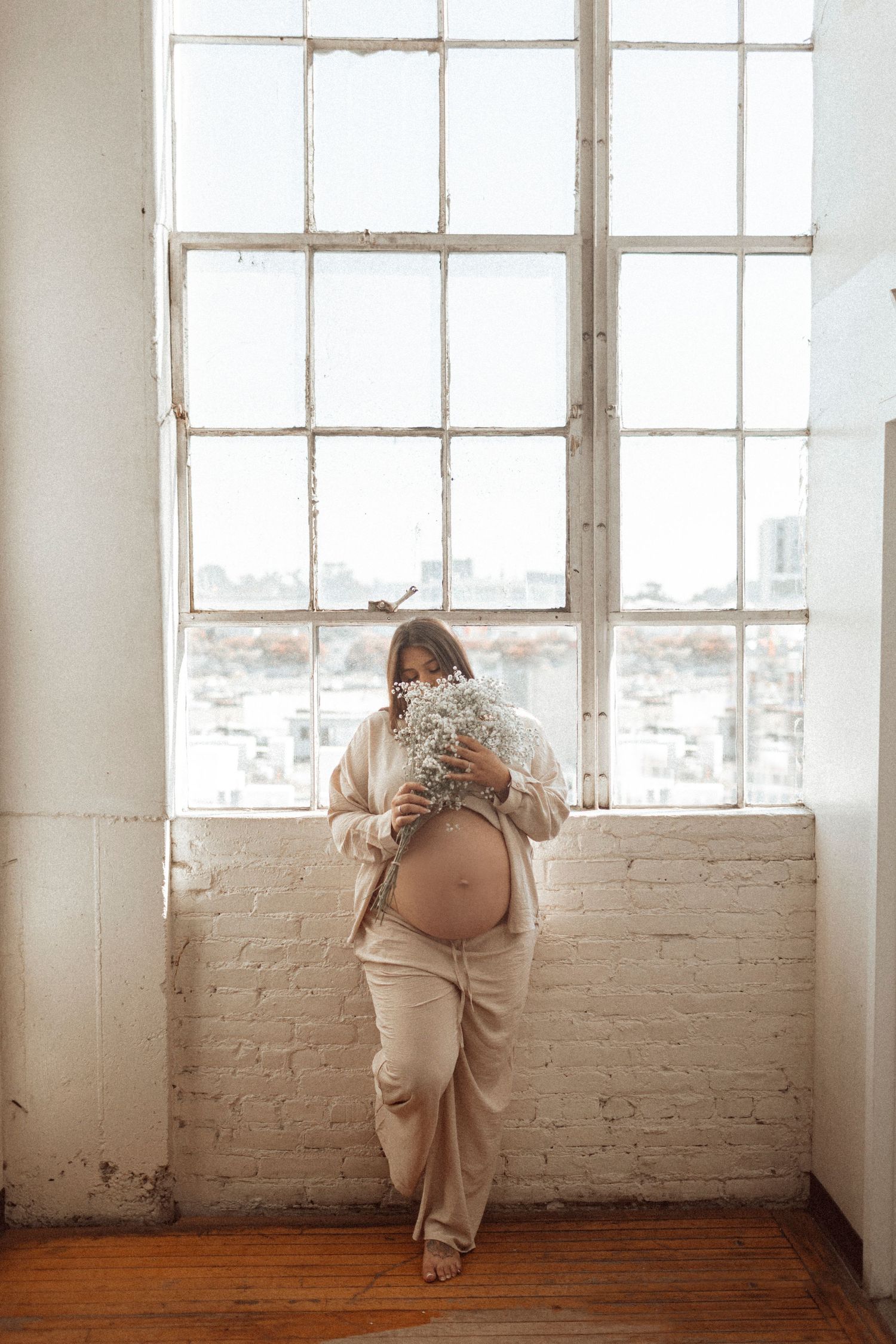 Maternity session in a studio with a brick wall, featuring neutrals, an exposed belly, and the woman holding a bouquet of flowers, creating