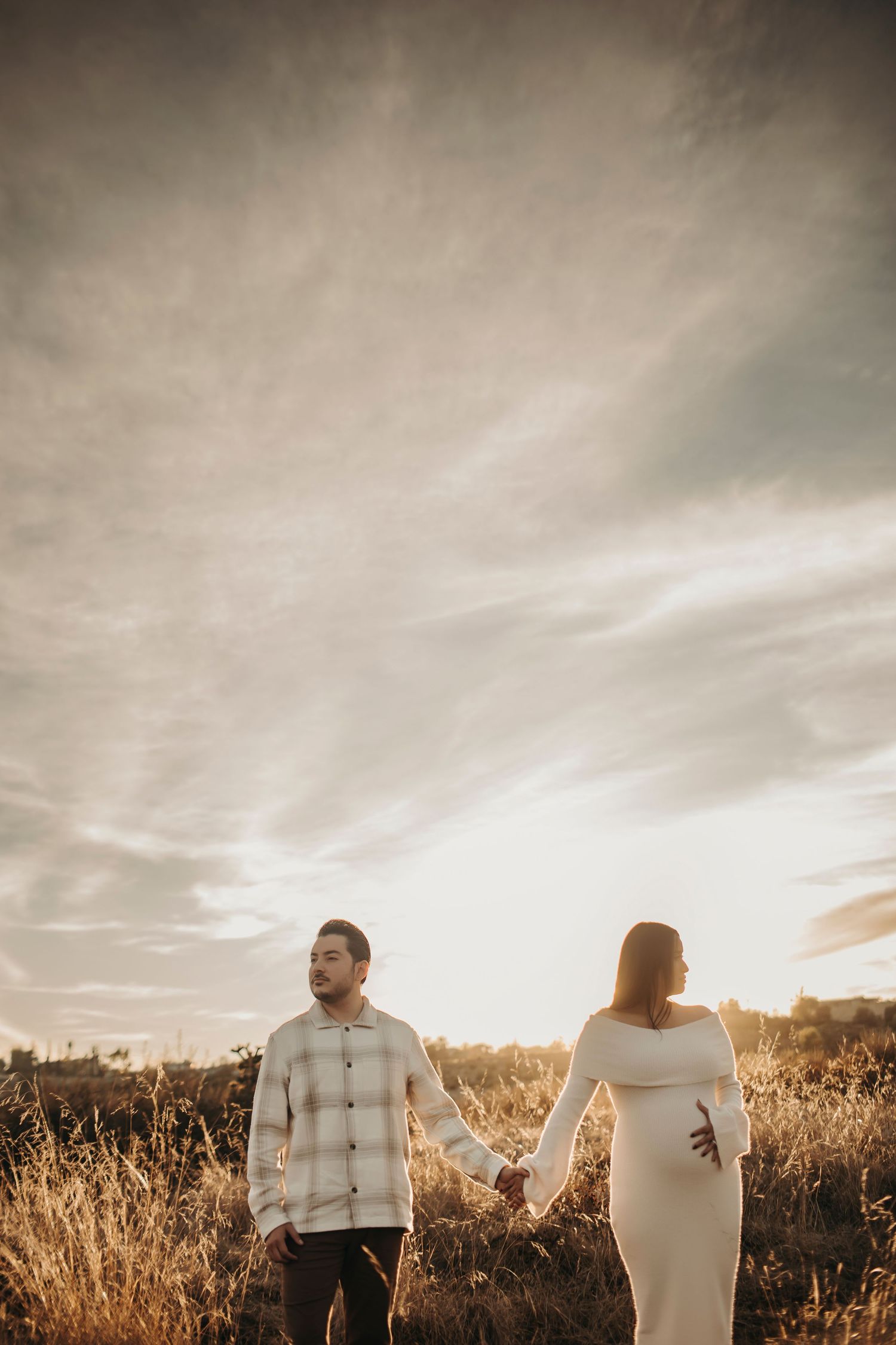 Maternity session in a golden field with a couple holding hands, both looking serious and facing opposite directions in dramatic lighting.