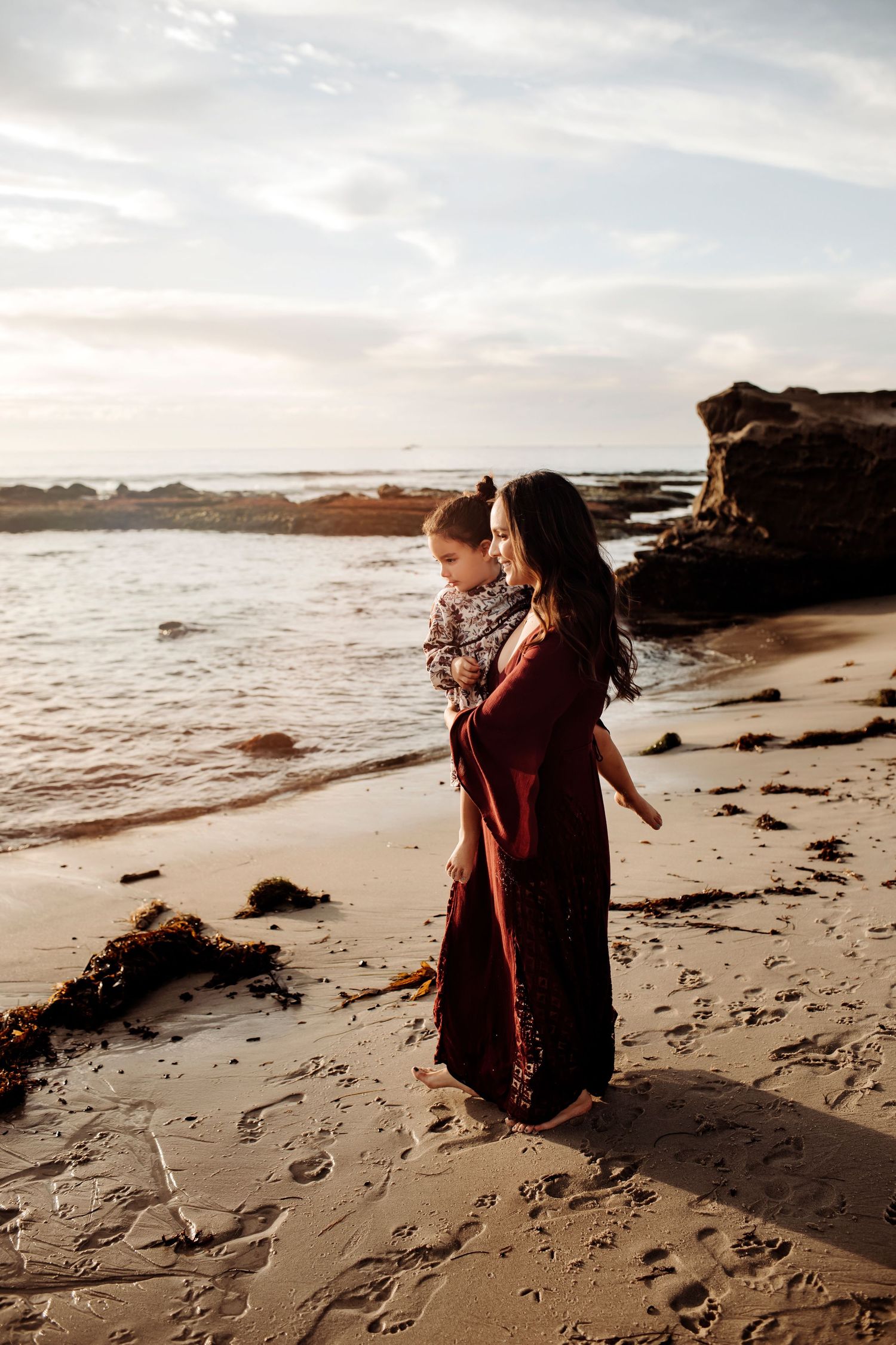 Maternity session on the beach, with the mother holding her toddler while walking along the shoreline, creating a joyful and serene moment.