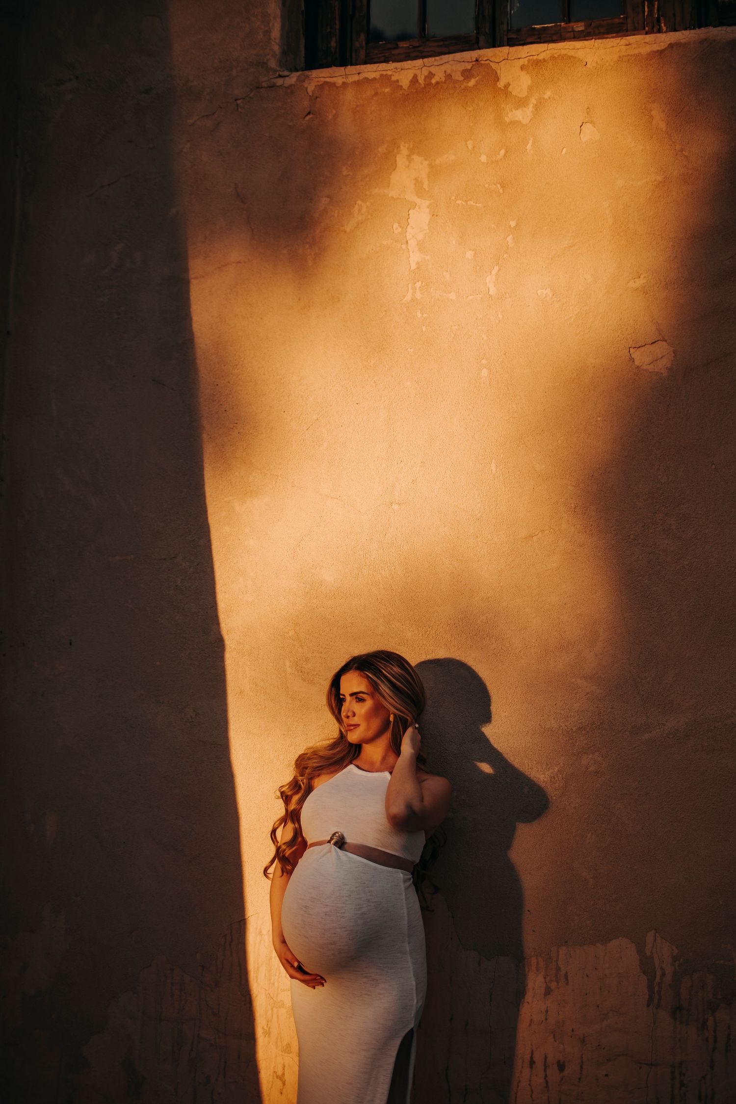 Maternity session featuring a woman in a white tight dress with long hair, holding her hair under harsh artistic light, creating a dramatic