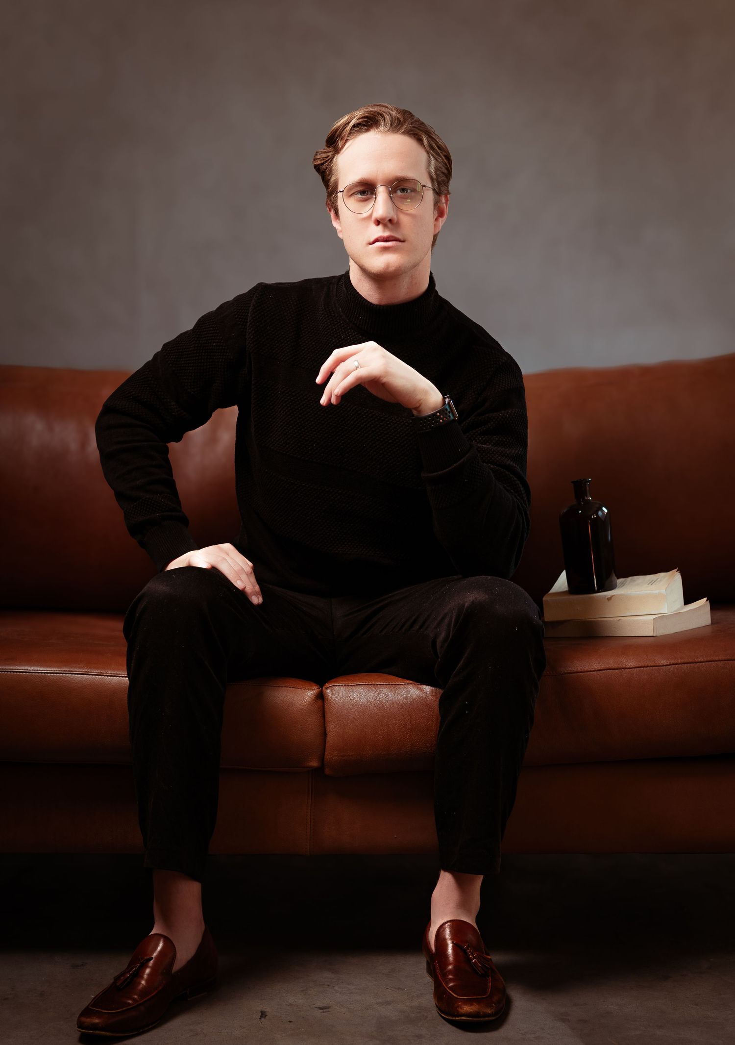 A person in a black turtleneck and pants sits confidently on a brown leather couch in a studio setting.