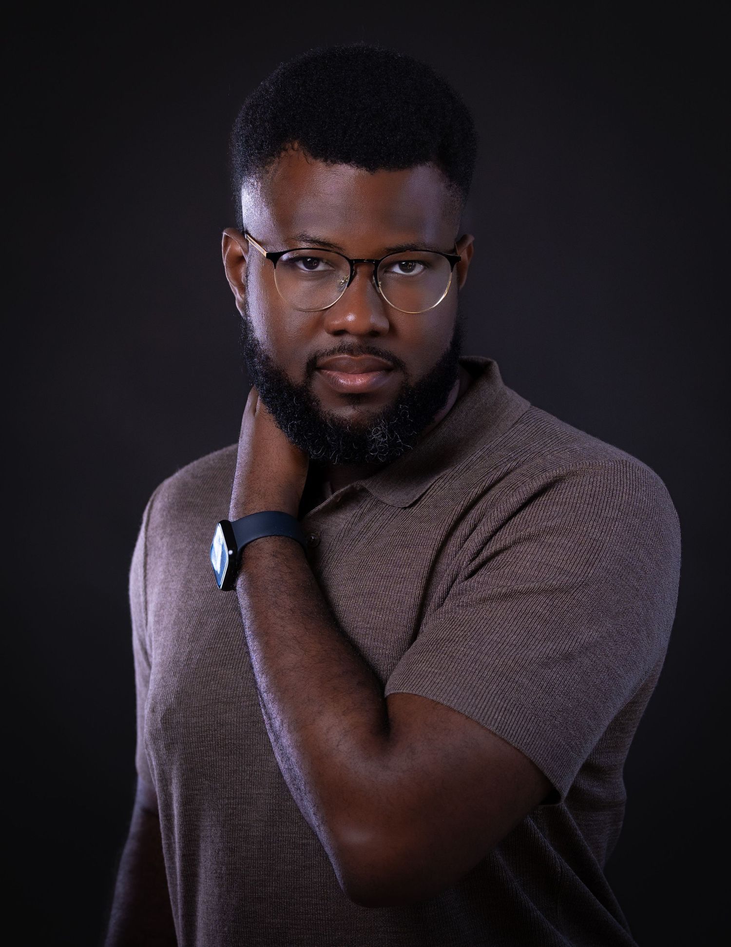 Studio portrait of a person in glasses and brown polo shirt posing with hand on neck against dark background.