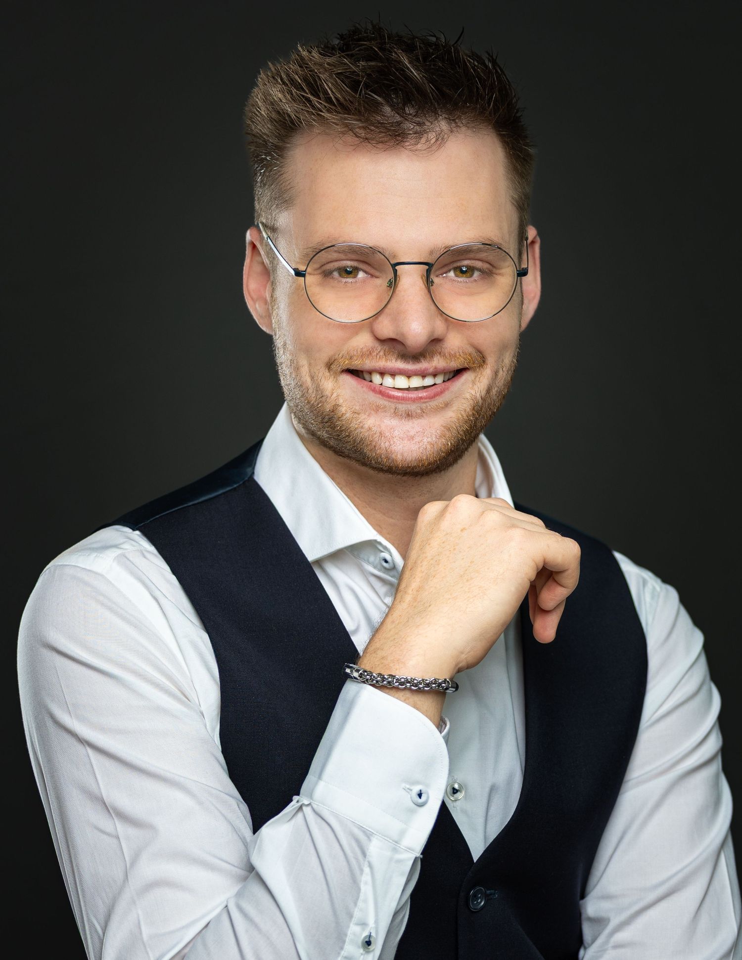 Professional headshot of person wearing glasses, white shirt and dark vest against dark background.