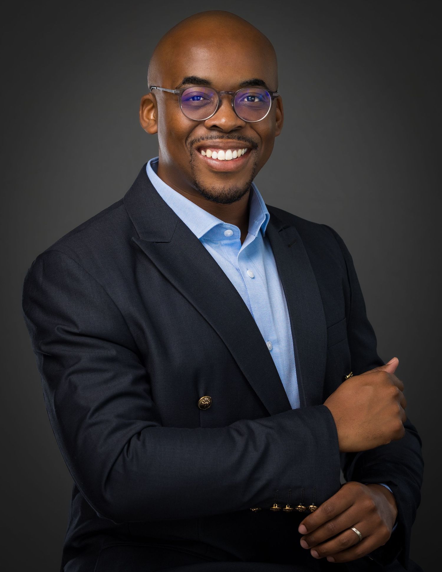 Professional headshot of person in navy blazer and blue dress shirt with purple glasses against dark background.