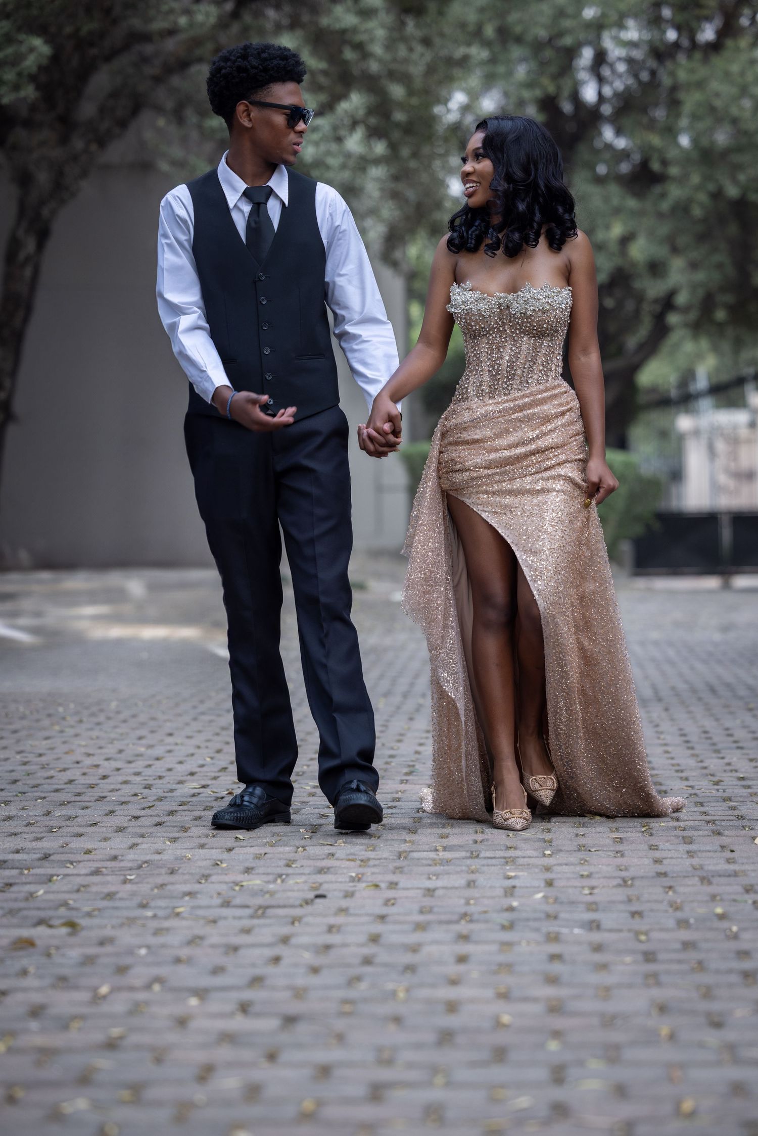 Two people in formal attire standing together on a cobblestone path, with one wearing an elegant champagne gown.