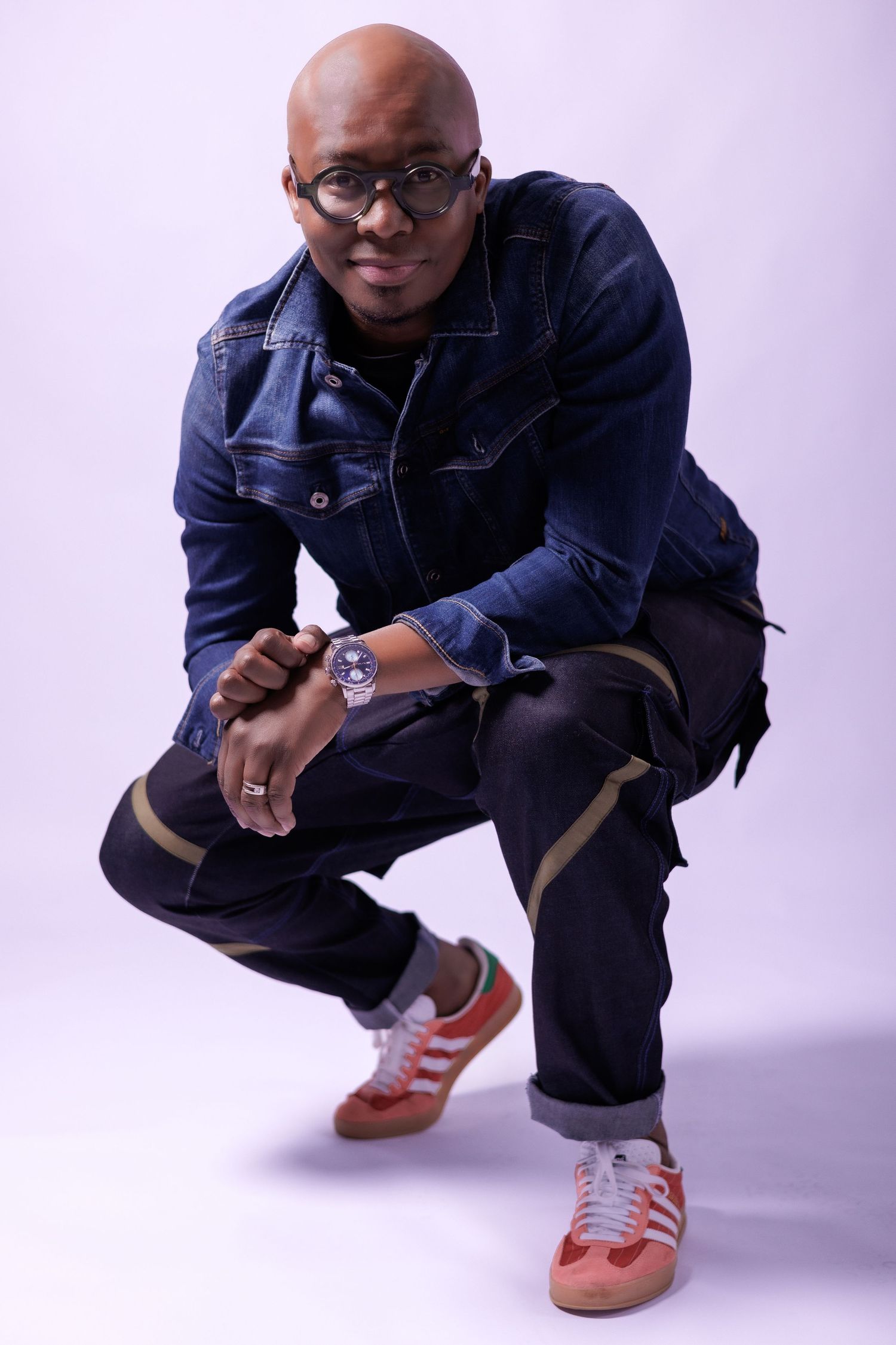 Stylish portrait of person in denim jacket and sneakers crouching against light background.