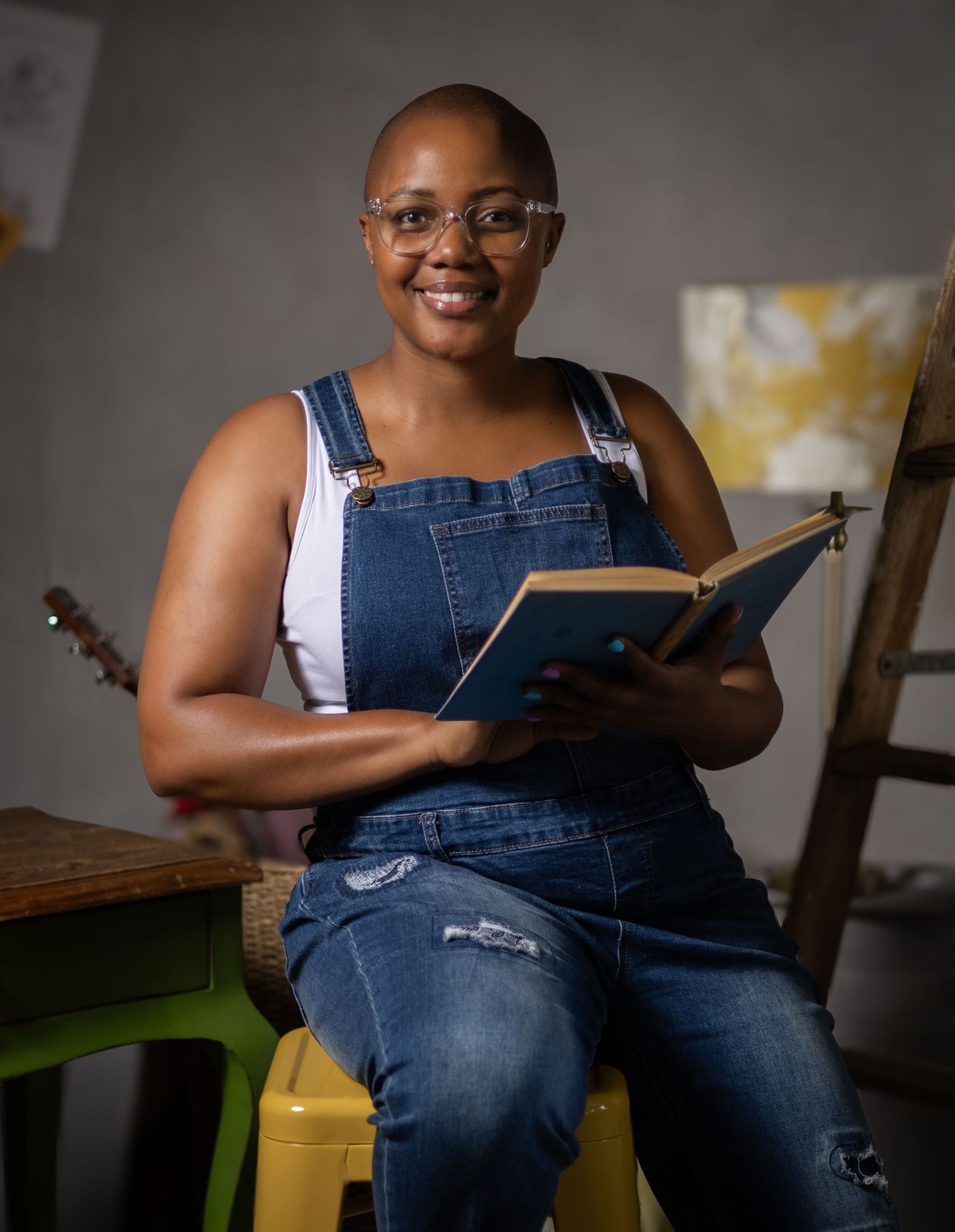 Person in denim overalls sits casually while holding a book in a creative artistic space with paintings visible behind them.