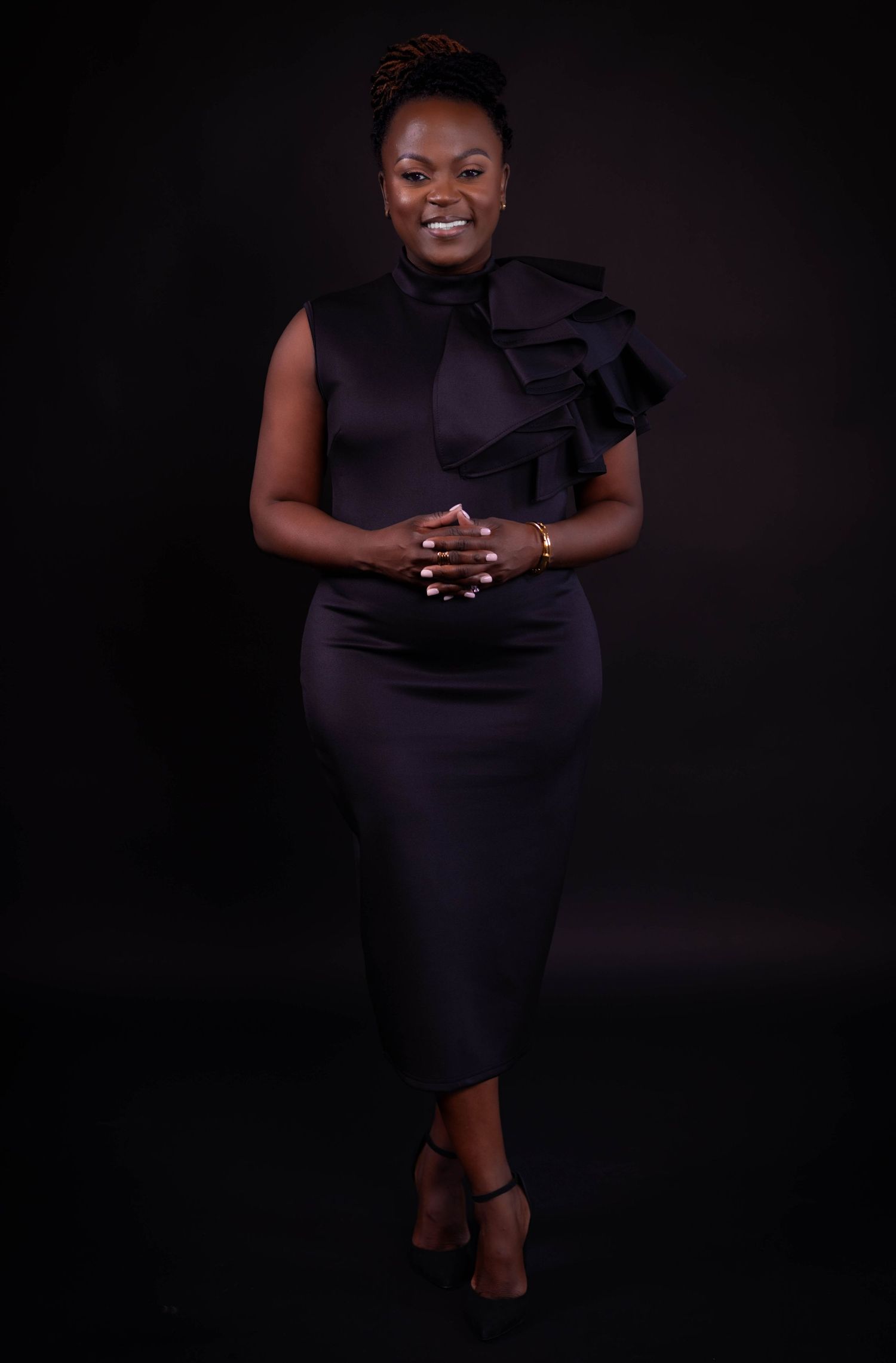 Elegant portrait of person in black fitted dress with ruffled shoulder detail against dark backdrop.