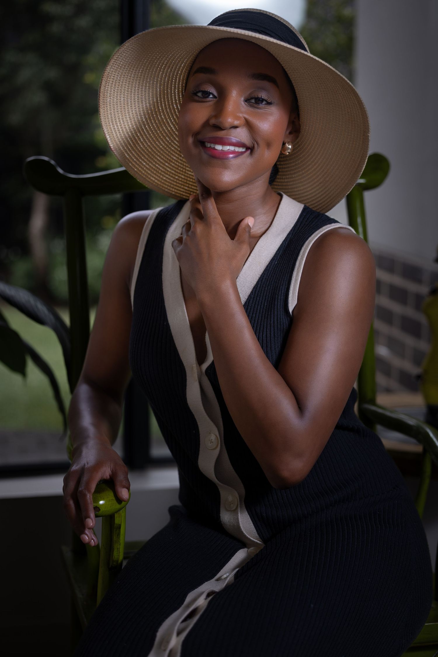 Elegant portrait of someone wearing a wide-brimmed sun hat and black dress with beige stripes sitting near plants.