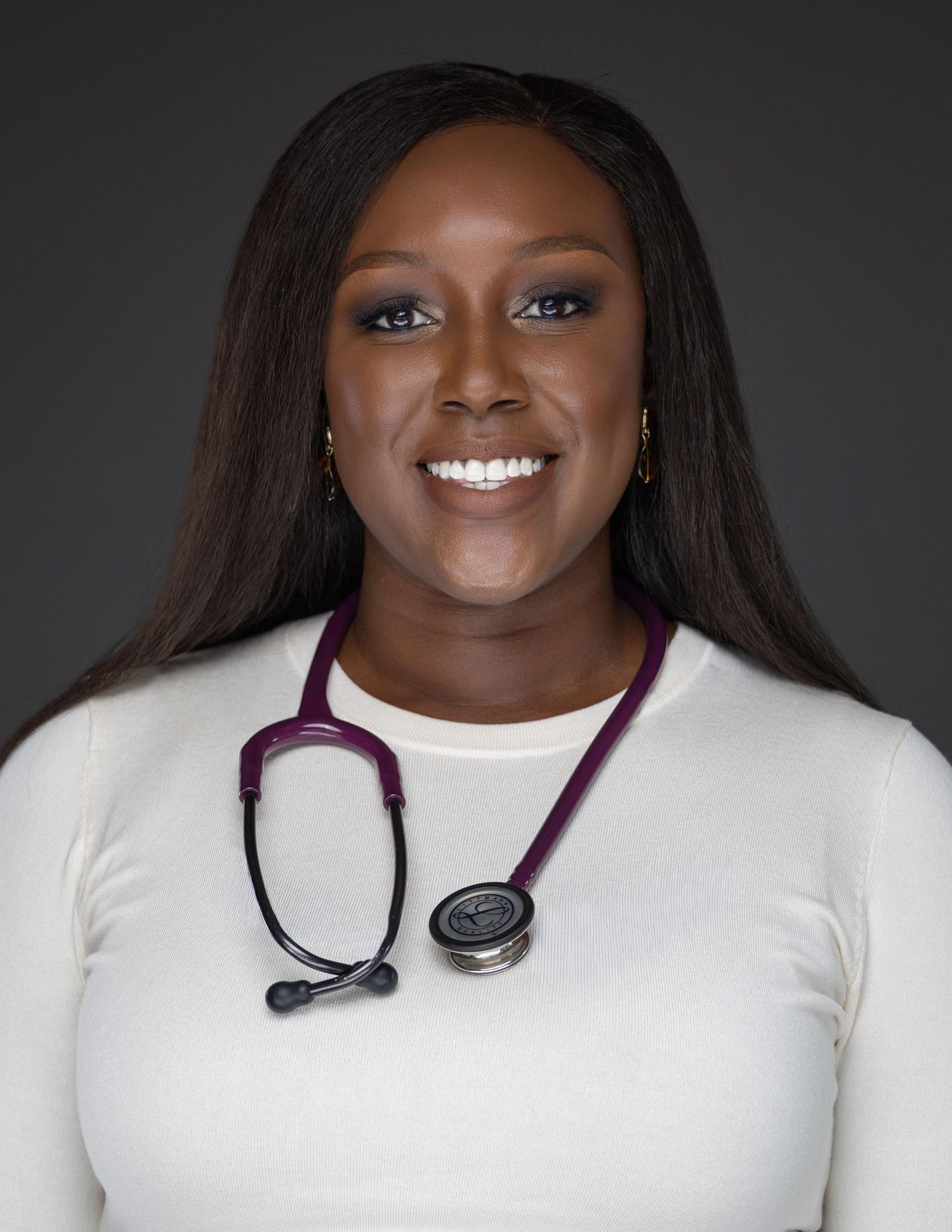 Healthcare professional wearing a white top and stethoscope smiles warmly against dark background.