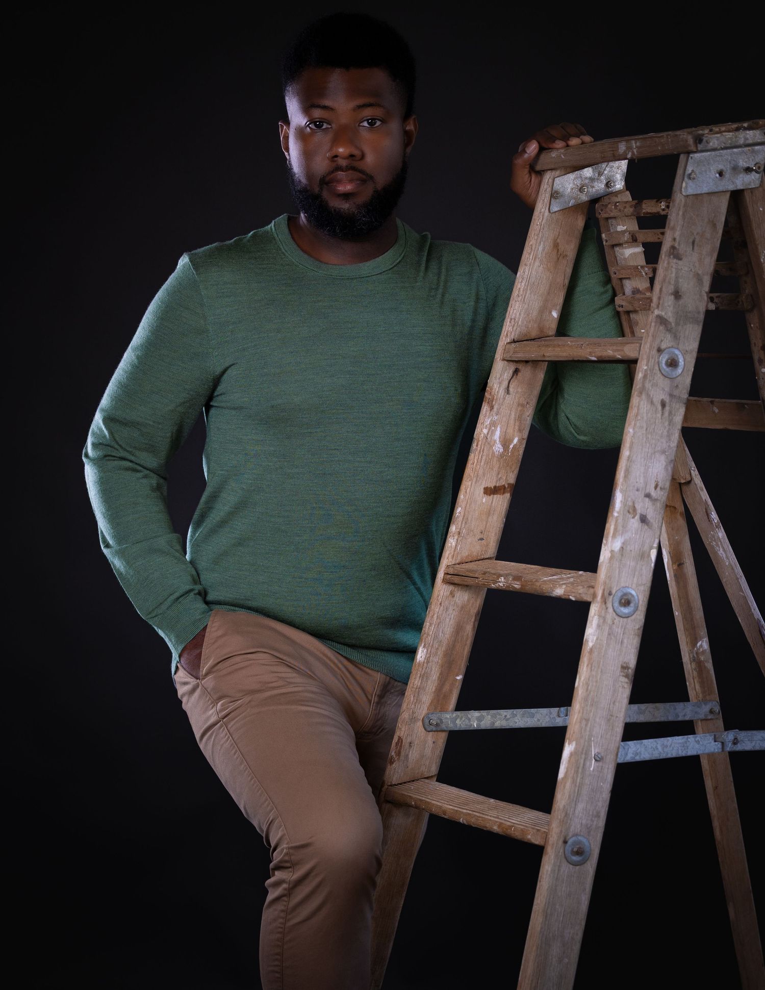 A person in a green sweater and khaki pants sits on a wooden ladder against a dark background.