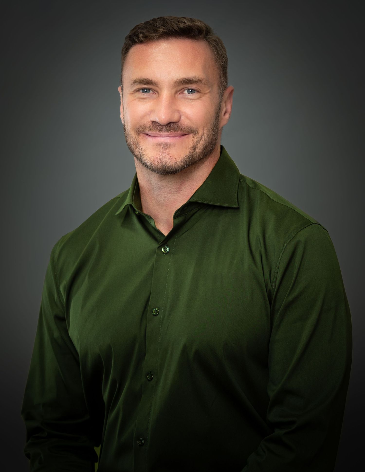Professional headshot portrait of a smiling person wearing a dark green collared shirt against a gray backdrop.