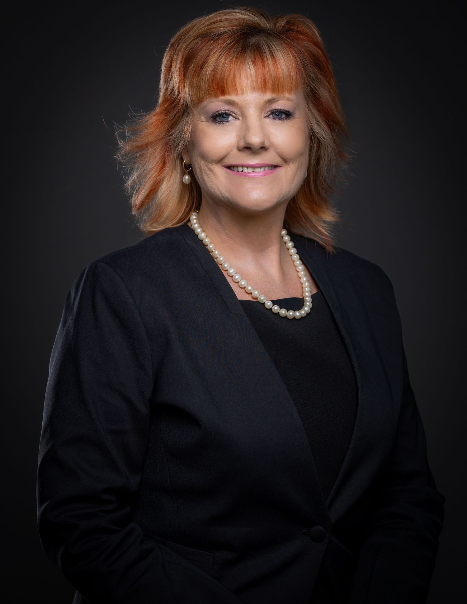 Professional business portrait of a person in a black blazer and pearl necklace against a dark background.