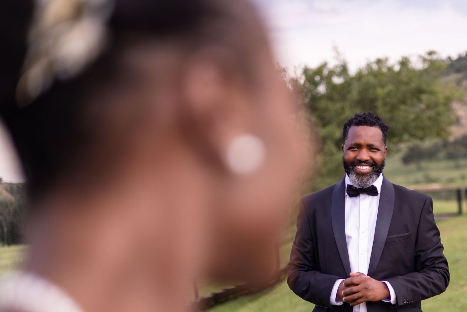 A happy groom in a black tuxedo and bow tie smiles during an outdoor wedding ceremony.